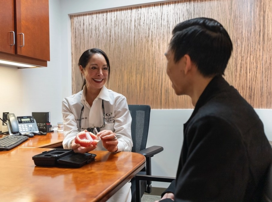 Dentist sitting at a table, explaining Invisalign treatment to a patient while holding a dental model and clear aligners