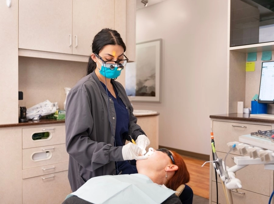 A dental professional wearing protective eyewear, a mask, and gloves is performing a procedure on a patient lying in a dental chair