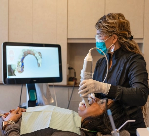 Dental hygienist using an intraoral camera to examine a patient's mouth, displayed on a nearby screen, with the patient seated in the dental chair.