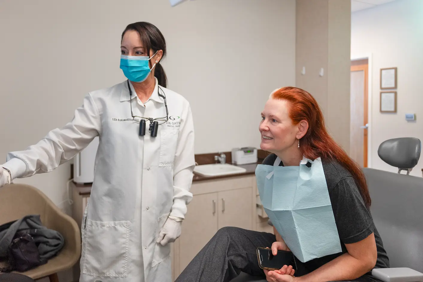 Dental hygienist using an intraoral camera to examine a patient's mouth, displayed on a nearby screen, with the patient seated in the dental chair.